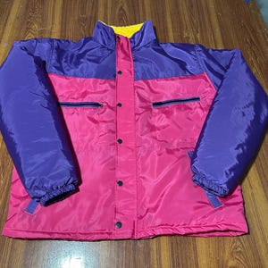 May include: A colour-blocked puffer jacket featuring a bright pink main body with purple sleeves and upper chest panels. The jacket includes a high collar with a yellow interior lining, a front snap-button closure, and two horizontal zippered chest pockets.
