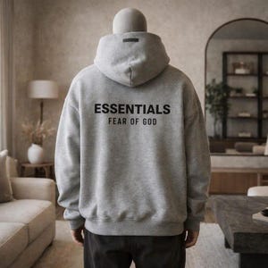 Op de afbeelding: Een grijze hoodie met het woord "ESSENTIALS" in zwarte letters op de rug, met daaronder "FEAR OF GOD". De hoodie heeft een capuchon en een klein zwart rechthoekje op de capuchon. De persoon draagt een donkere broek.