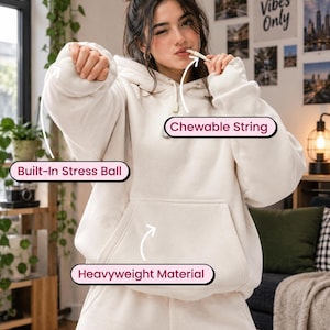 May include: A cream-colored hoodie featuring a built-in stress ball in the sleeve, a chewable string at the neckline, and a front kangaroo pocket made of heavyweight material. The garment includes labels pointing to these specific functional features.