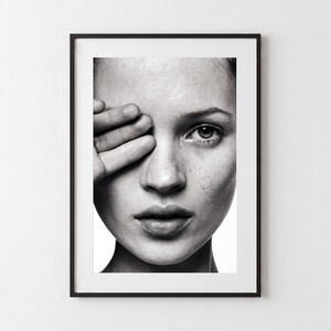 May include: A black and white framed portrait print featuring a close-up of a face with one hand partially covering the eye. The image captures detailed skin texture, a single visible eye, and a neutral expression within a thin black frame.