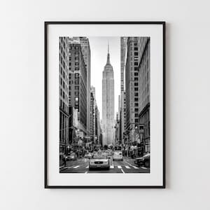 May include: A black and white framed wall art print featuring a vertical perspective of a New York City street. The Empire State Building stands tall in the center, framed by towering skyscrapers on both sides, with cars driving on the road.