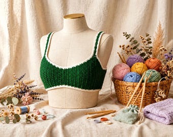 May include: A dark green crochet crop top with white trim on the straps and neckline, displayed on a mannequin. The top features a ribbed texture and is positioned near a wicker basket filled with colorful yarn balls, wooden hooks, and dried flowers.
