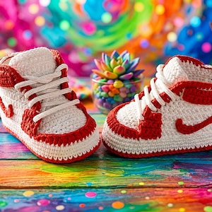 Op de afbeelding: Een paar gehaakte wit met rode high-top sneakers met witte veters, een hoge enkelkraag en een rood, swoosh-achtig detail aan de zijkant. De schoenen zijn gemaakt met een gestructureerde garensteek die het uiterlijk van sportschoenen nabootst.