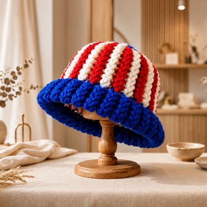 May include: A chunky knit bucket hat featuring vertical stripes in alternating red and white yarn, finished with a wide, solid blue ribbed brim. The hat is constructed from thick, textured yarn and displays a distinct, hand-crafted appearance.