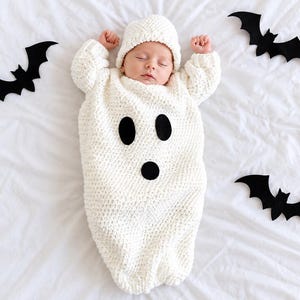 Può includere: Un sacco nanna per neonati in maglia bianca con berretto abbinato, caratterizzato da due occhi ovali neri e una bocca circolare nera per assomigliare a un fantasma. Tre pipistrelli di carta nera sono posizionati attorno al sacco su un tessuto.