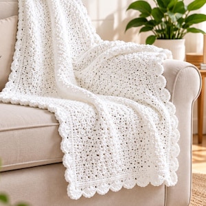 May include: A white crocheted blanket with a textured pattern and a scalloped edge draped over a beige sofa. A throw pillow on the sofa features the text Home Sweet Home with a heart symbol, and a wooden sign nearby reads Good Things Take Time.