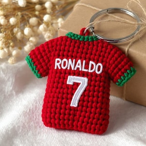Puede incluir: Un llavero de ganchillo rojo con forma de camiseta de fútbol con ribetes verdes en el cuello y las mangas. La parte posterior presenta el texto bordado en blanco RONALDO sobre el número 7 en blanco, unido a una anilla de metal plateado.
