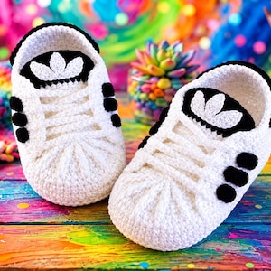 Op de afbeelding: Een paar wit gehaakte babyschoentjes in de vorm van sneakers. De schoenen hebben zwarte gehaakte accenten op de tong, hiel en zijkanten, met een wit klaverblad-embleem op de tong en witte gehaakte veters over de bovenkant van de schoen.