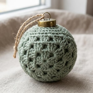 May include: A spherical ornament covered in sage green crochet yarn featuring a classic granny square pattern. The top of the ornament is finished with a metallic bronze cap and a natural jute twine loop for hanging, creating a textured appearance.