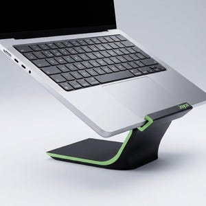 Puede incluir: Un portátil plateado con teclado negro descansa sobre un soporte curvo de color negro y verde lima. La palabra Joyce está impresa en verde lima en el borde frontal del soporte. El portátil está inclinado hacia arriba sobre la base.