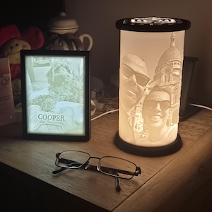 May include: A rectangular light box displaying a dog with a bone-shaped tag labeled COOPER and a cylindrical lithophane lamp featuring a portrait of two people wearing sunglasses in front of the Sacré-Cœur Basilica, with black-framed eyeglasses.