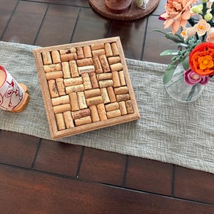 May include: A wooden tray filled with wine corks in a mosaic pattern. A coffee mug with a cartoon character design sits on a coaster to the left. A vase of colorful flowers is on the right, all on a grey table runner.
