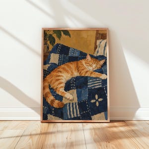 May include: A framed art print featuring an orange tabby cat sleeping on a blue and white patchwork quilt. The quilt displays various geometric patterns and shades of indigo. The cat is curled up in the centre, resting on the textured fabric.