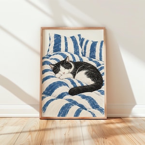 May include: A framed rectangular print featuring a black and white cat curled up asleep on a bed with blue and white striped bedding and pillows. The cat has a black back and head with a white face and paws, resting on the textured fabric surface.