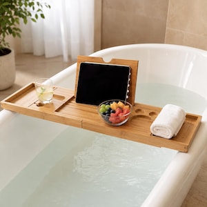 Peut inclure: Un plateau de baignoire rectangulaire en bambou doté d'un support central réglable pour tablette, d'une découpe circulaire pour un verre, d'un petit bol de fruits mélangés et d'une serviette blanche roulée sur le côté droit, posé sur une baignoire.