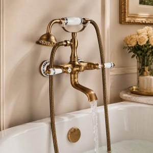 May include: A wall-mounted bathtub faucet set in an antique brass finish with white ceramic accents featuring blue floral patterns. The set includes a handheld shower head, a curved spout with flowing water, and two cross-style handles marked H and C.