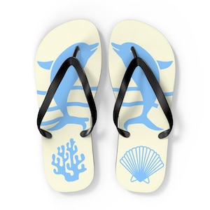 Op de afbeelding: Een paar crèmekleurige slippers met zwarte rubberen bandjes. De linkersandaal toont een lichtblauwe dolfijnafbeelding boven een koraalrif-icoon, terwijl de rechtersandaal een lichtblauwe dolfijnafbeelding boven een schelp-icoon toont.