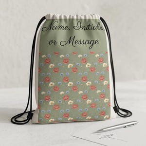 May include: A drawstring backpack featuring a sage green background with a repeating floral pattern of red, white, and blue flowers. The upper portion of the bag displays the black text Name, Initials, or Message in a cursive script font.