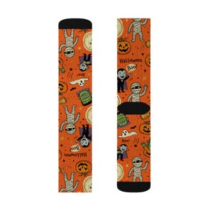 May include: A pair of orange socks featuring a repeating Halloween pattern of mummies, pumpkins, ghosts, and vampires. The design includes the text "Halloween" and "Boo!" scattered across the fabric, with black cuffs at the top and bottom.
