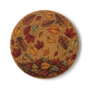 May include: A circular decorative plate featuring a detailed illustration of swirling autumn leaves in shades of red, orange, yellow, and brown. The design includes various leaf shapes and small acorns scattered across a warm tan background.