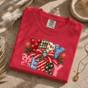Puede incluir: Una camiseta roja de cuello redondo con un gráfico que incluye el texto VERY MERRY. Las letras están rellenas con varios patrones navideños como cuadros, copos de nieve y rayas, decoradas con un lazo rojo, ramas de pino y estrellas.