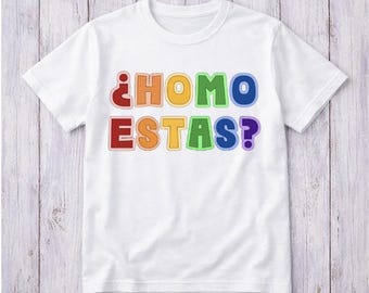 May include: White t-shirt with the text "¿HOMO ESTAS?" in rainbow-coloured letters. The text is outlined in a darker colour, and the shirt has a crew neck and short sleeves. The shirt is displayed on a light wood background.