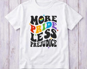 May include: A white short-sleeved t-shirt featuring the text "MORE PRIDE LESS PREJUDICE" in a bold, wavy black font. The word "PRIDE" is multicoloured, and a small rainbow-coloured heart sits above the text, surrounded by tiny star icons.