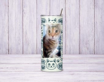 May include: A cylindrical tumbler with a metal straw featuring a central photographic portrait of a brown tabby cat. The background of the tumbler is light-coloured and decorated with dark grey line art of cat faces, paw prints, and the word MEOW.