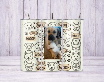 May include: A cylindrical tumbler with a cream-coloured background featuring a repeating pattern of black line-art dog faces, paw prints, bones, and the word WOOF in speech bubbles. A rectangular photograph of a tan dog holding a toy is centred.