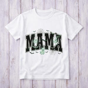 May include: A white short-sleeved t-shirt featuring the word MAMA in large black block letters. The design includes pale green ghost illustrations, black spiderwebs, and small black bats scattered around and within the lettering.
