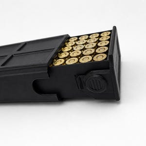 May include: A black rectangular magazine filled with brass-colored bullets. The magazine has a textured surface and a sliding top. The bullets are neatly arranged inside the magazine, which is set against a white background.