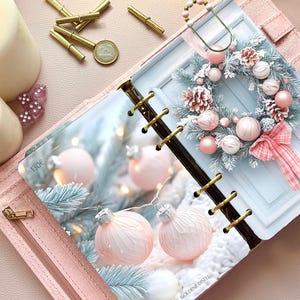 Könnte beinhalten: Ein hellrosa Planer mit Ringbuchmechanik, der eine Seite mit dem Druck eines Miniaturkranzes enthält, verziert mit rosa Ornamenten und einer karierten Schleife, sowie zwei hängenden rosa Kugeln und einem verschneiten Tannenzweigmotiv.