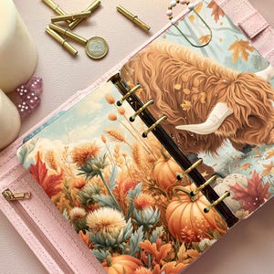 Könnte beinhalten: Ein hellrosa Ringplaner mit einem illustrierten Einleger, der ein zotteliges Hochlandrind in einer herbstlichen Landschaft mit orangefarbenen Kürbissen, goldenem Weizen und herbstlichem Laub zeigt. Der Planer hat eine goldfarbene Ringmechanik.