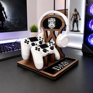 Könnte beinhalten: Eine Gaming-Station aus Holz mit Kopfhörerhalterung, zwei weißen Gamecontrollern und einem Tablett. Der Ständer trägt den Text "GAMER DAD" und "I LOVE YOU DAD". Das Headset und die Controller sind weiß.