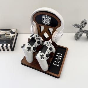 Può includere: Una stazione di gioco in legno con cuffie bianche, due controller bianchi e un supporto per telefono. Il supporto per le cuffie ha la scritta "GAMER DAD" e "I LOVE YOU DAD" è sul supporto per telefono. La stazione è marrone su sfondo bianco.