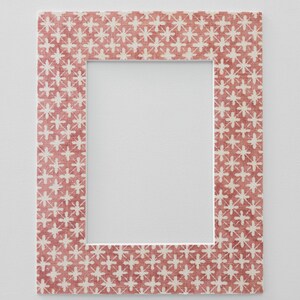May include: A rectangular picture frame featuring a repeating pattern of white cross-like shapes on a muted red background. The frame has a flat, wide border with a visible fabric-like texture, surrounding a central rectangular opening for a photo.