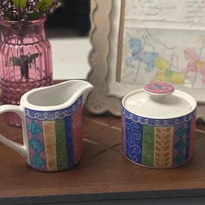 May include: A white ceramic creamer and a matching lidded sugar bowl featuring a colorful vertical panel pattern in shades of blue, green, pink, and tan with decorative swirls, diamond shapes, and leaf motifs around the exterior surfaces.