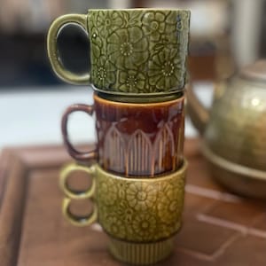Puede incluir: Una pila vertical de tres tazas de cerámica. La taza superior es de color verde oliva con un patrón floral en relieve. La taza central es de color marrón brillante con detalles acanalados verticales. La taza inferior es verde oliva claro.