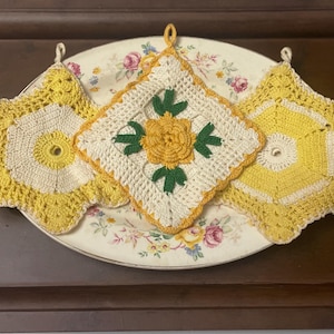 May include: Three handmade crochet potholders in yellow and cream yarn. Two are star-shaped with central circular openings, and one is a square featuring a central yellow flower with green leaves. Each piece includes a small loop for hanging.