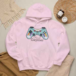 Peut inclure: Un sweat-shirt à capuche rose clair avec un motif de manette de jeu vidéo bleu clair arborant un visage souriant, des cœurs rouges et des arcs-en-ciel sur les poignées. Le texte "Proud Gaymer" est imprimé en noir sous le motif.