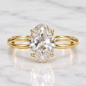 Könnte beinhalten: Ein Goldring mit einem großen, oval geschliffenen, klaren facettierten Edelstein, der von vier Krappen gehalten wird. Die Ringschiene besteht aus poliertem Gelbgold und teilt sich zu beiden Seiten des Steins in zwei feine, verschlungene Bögen.