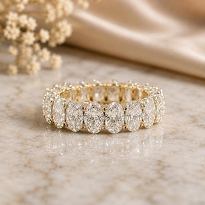 May include: An eternity band ring crafted from yellow gold, featuring a continuous row of sparkling oval-cut clear gemstones set in a prong setting that encircles the entire circumference of the band, creating a uniform and brilliant appearance.