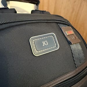May include: A black ballistic nylon backpack featuring a top carry handle with brown leather trim, multiple zippered compartments, and a rectangular patch on the front centre displaying the embossed silver-toned initials JG in a serif font.