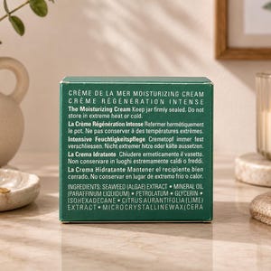 La Mer Crème de La Mer miniature cream box photographed on a marble surface with decorative vase, books, and candle in a modern home setting.