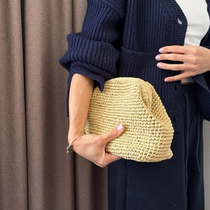 以下が含まれることがあります： 明るいタン色の織りストロークラッチ。ネイビーブルーのカーディガンと黒いパンツを着た人がクラッチを持っています。背景は茶色のカーテンです。
