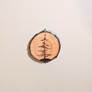 May include: A wooden round slice with a burnt design of a pine tree on it.