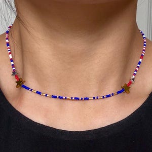May include: A beaded choker necklace featuring a repeating pattern of small red, white, and blue beads. The design includes metallic gold-toned star charms and small gold-colored beads interspersed along the lower section of the strand.