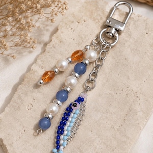 May include: A silver-toned metal keychain featuring a lobster clasp and a dangling charm shaped like a fish made of small blue, light blue, and white beads. The keychain also includes a strand of white pearls, blue beads, and amber-colored beads.