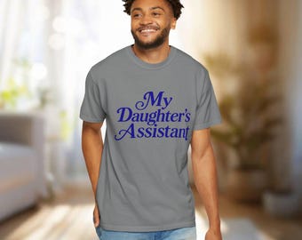 May include: A grey short-sleeved t-shirt featuring the text My Daughter's Assistant printed in dark blue lettering across the chest. The shirt has a crew neckline and a standard fit, providing a simple and casual appearance for everyday wear.