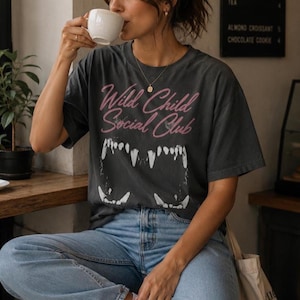 May include: Dark grey t-shirt with the text "Wild Child Social Club" in pink script above a white graphic of a toothy grin. The person is wearing blue jeans and drinking from a white mug.